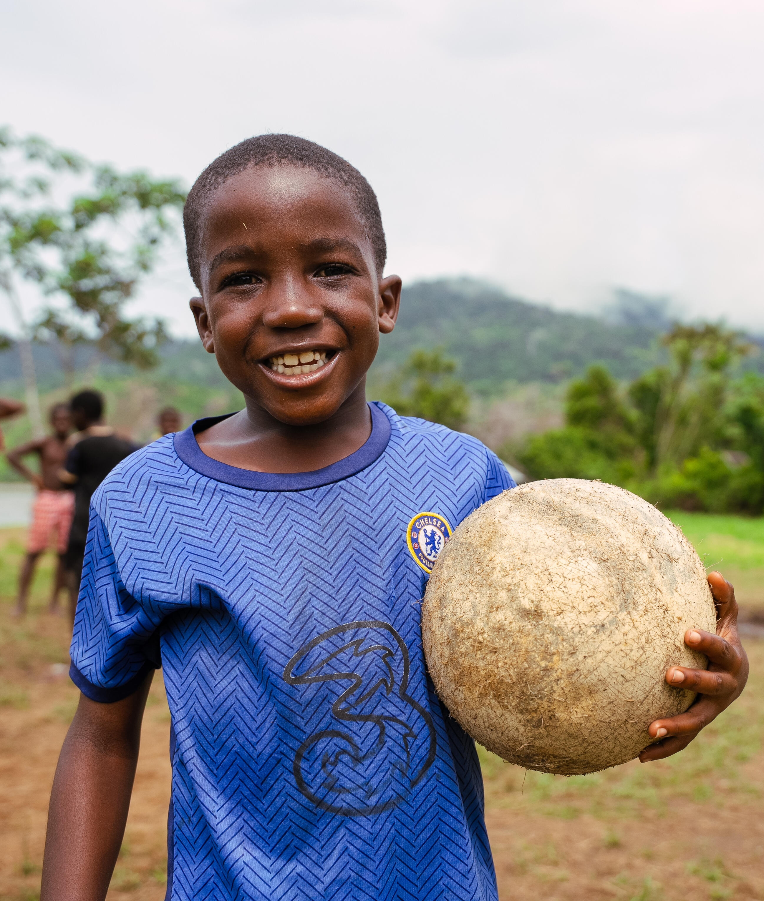 Un niño de Surinam lleva la equipación del Chelsea FC y sostiene un balón de fútbol. Sonríe a la cámara. Detrás de él se ven unas colinas verdes y un grupo de niños.